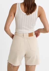 Pantalones cortos de mezclilla beige claro con dobladillo sin acabar y bolsillos traseros, combinados con una camiseta de tirantes blanca con un patrón texturizado.