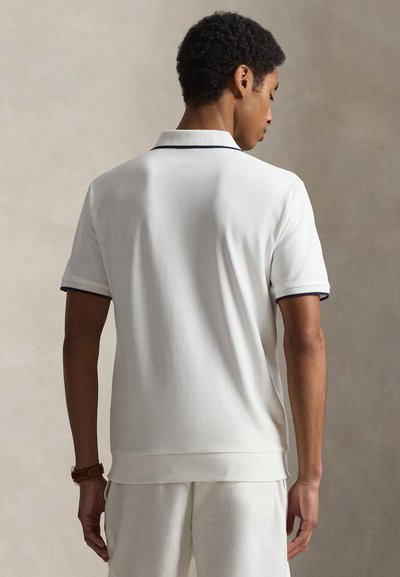 Homme aux cheveux courts et bouclés, portant un polo blanc à manches courtes avec un liseré foncé sur le col et les manches, face à un fond neutre, de dos.