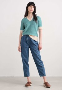 Neule turquoise-värissä, V-kaula-aukolla ja kermanvärisillä yksityiskohdilla, yhdistettynä vaaleansinisiin naru-/denim-housuihin ja ruskeisiin puukengät.