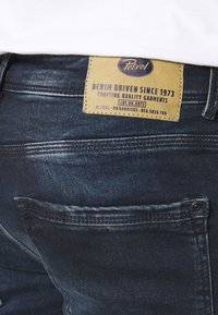 Siniset denim-farkut, joissa on vaaleanruskea nahkalappu, jossa on kohokuvioitu teksti, viiden taskun muotoilu ja hienovarainen kuluminen kankaassa.
