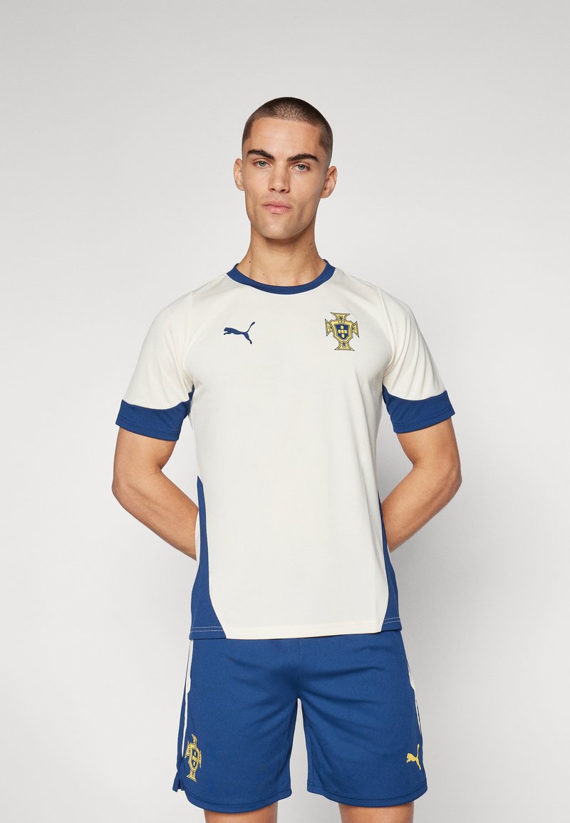 Maglietta sportiva bianca con accenti blu, dotata di maniche corte e scollo rotondo. Presenta un logo sul petto ed è abbinata a pantaloni blu.
