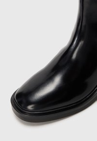 Bota negra de cuero brillante hasta el tobillo con puntera cuadrada redondeada y suela gruesa negra sobre un fondo blanco liso.