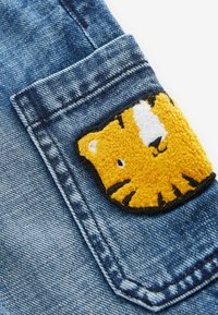 Tissu en denim avec une poche ornée d'un patch en forme de tête de tigre jaune texturée, avec des rayures noires et des détails blancs.
