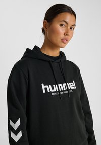 Młoda kobieta z ciemnymi włosami w czarnej bluzie "hummel" z białym napisem i wzorem w chevron na rękawie, na jednolitym tle.