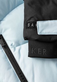 Ovratnik crnog puf jakne ističe "BAKER" brendiranje. Pozadina je svijetloplava tkanina, s crnim patentnim zatvaračem i kontrastnim oznakama.