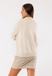 Maglione beige a maglia con colletto e orlo a coste, caratterizzato da una vestibilità comoda. Indossato sopra una gonna a quadri, che evidenzia il tessuto testurizzato e le cuciture.