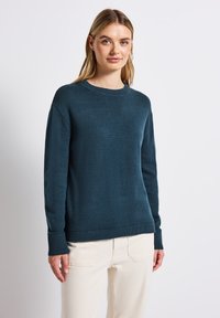Türkiser Strickpullover mit runden Halsausschnitt, gerippten Bündchen und Saum. Getragen mit cremefarbenen Hosen vor einem schlichten weißen Hintergrund.