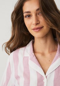 Chemise légère en lin avec rayures verticales roses et blanches, col court et fermeture à boutons sur le devant. Texture douce et coupe décontractée.