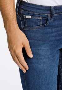 Blaue Jeans aus Denim mit schmaler Passform, klassischem Fünf-Taschen-Design und sichtbaren Nähten. Der Stoff hat eine glatte Textur.