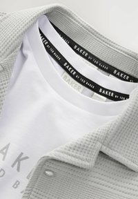 Grå tekstureret jakke over en hvid T-shirt med et trykt logo. Etiketterne viser "BAKER by TED BAKER" i sort og hvid.
