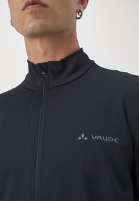 Czarna bluzka zapinana na zamek, wykonana z teksturowanej tkaniny, z wysokim kołnierzem i subtelnym szarym logo "VAUDE" po lewej stronie.