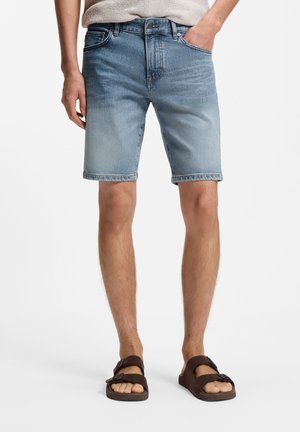 Man wearing light blue denim shorts and dark brown sandals, standing with one hand in pocket against a white background.