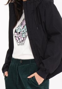 Giacca nera con cappuccio e zip frontale, indossata sopra una maglietta bianca con grafiche colorate e design astratti, abbinata a pantaloni in velluto a coste verdi.