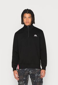 Felpa nera con zip e colletto alto, presenta un piccolo logo bianco sul petto e un'etichetta rossa a contrasto sull'orlo. Indossata con pantaloni cargo mimetici.