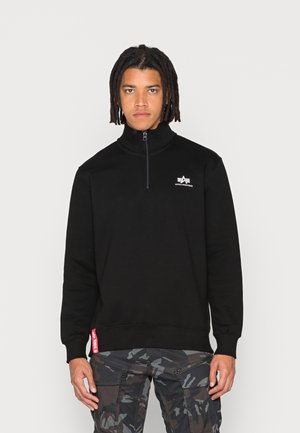 Black zip-up sweatshirt with a high collar, features a small white logo on the chest and a red accent tag at the hem. Worn with camo cargo pants.