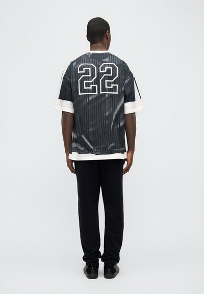 Άντρας που στέκεται με την πλάτη, φοράει αθλητική φανέλα με μαύρες και άσπρες ρίγες και αριθμό 22, μαύρο παντελόνι και μαύρα αθλητικά παπούτσια σε απλό φόντο.