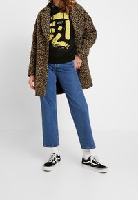 Abrigo de estampado de leopardo, sudadera negra con gráfico amarillo, vaqueros rectos azules, zapatillas en blanco y negro, exhibidos sobre un fondo neutro.