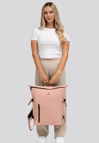 Donna con lunghi capelli biondi tiene uno zaino grande rosa con cerniere nere, in piedi su uno sfondo grigio uniforme.