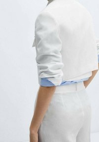 Valkoinen lyhyt takki käärittyine hihoineen vaaleansinisestä raidallisesta paidasta päälle. Sileä tekstuuri, minimalistinen muotoilu ja selkeä siluetti.