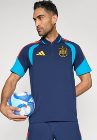 Tmavomodré športové tričko s krátkym rukávom s tyrkysovými akcentmi a červenými pruhmi, s golierom na zips a zlatým emblémom. Drží modro-bielu futbalovú loptu.