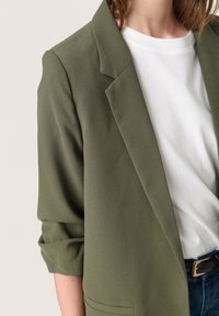 Olivově zelený blazer se strukturovaným designem, s klopami, ohrnutými rukávy a hladkou texturou, nošený přes bílý tričko.