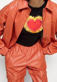 Giacca in ecopelle arancione e pantaloni abbinati, con una camicia nera decorata da un cuore colorato. Finitura testurizzata, design sportivo.
