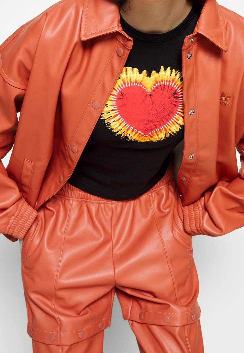 Giacca in ecopelle arancione e pantaloni abbinati, con una camicia nera decorata da un cuore colorato. Finitura testurizzata, design sportivo.
