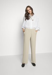 Calças largas beiges com cintura elástica e bolsos laterais, combinadas com uma camisa branca de botões e um lenço estampado. Sapatos pretos.