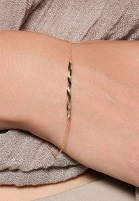 Złota bransoletka z wygiętym wzorem rurki z teksturowaną powierzchnią. Jest przymocowana delikatnym łańcuszkiem i noszona na jasnej karnacji skóry.