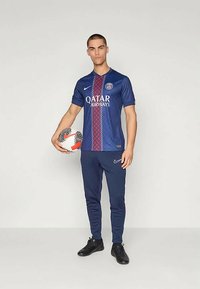 Tummansininen Nike-jalkapallovarustus, jossa on Qatar Airways -logo ja kuvioitu etuosa. Malli pitää jalkapalloa kädessään ja hänellä on päällään yhteensopivat housut ja kengät.