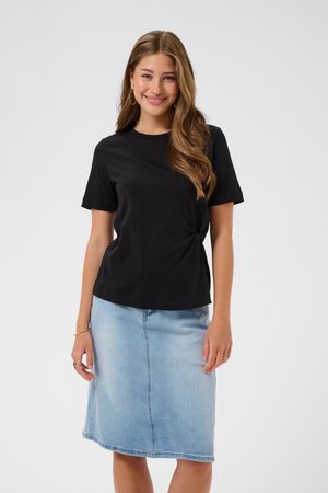 Lachende vrouw met lang golvend haar, gekleed in een zwart topje met korte mouwen en een lichtblauwe denim rok tot op de knie, staand tegen een effen achtergrond.