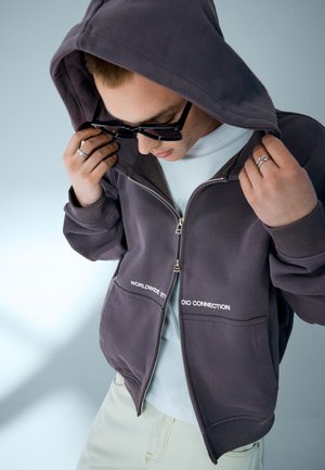 Osoba si upravuje kapucňu na tmavosivej mikine na zips s bielym textom, má na sebe slnečné okuliare, biele tričko a svetlé nohavice, stojí pred jednoduchým pozadím.