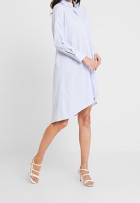 Šaty s modro-bílými pruhy, s knoflíky a dlouhými rukávy, se asymetrickým lemem, doplněné bílými sandály na vysokém podpatku.