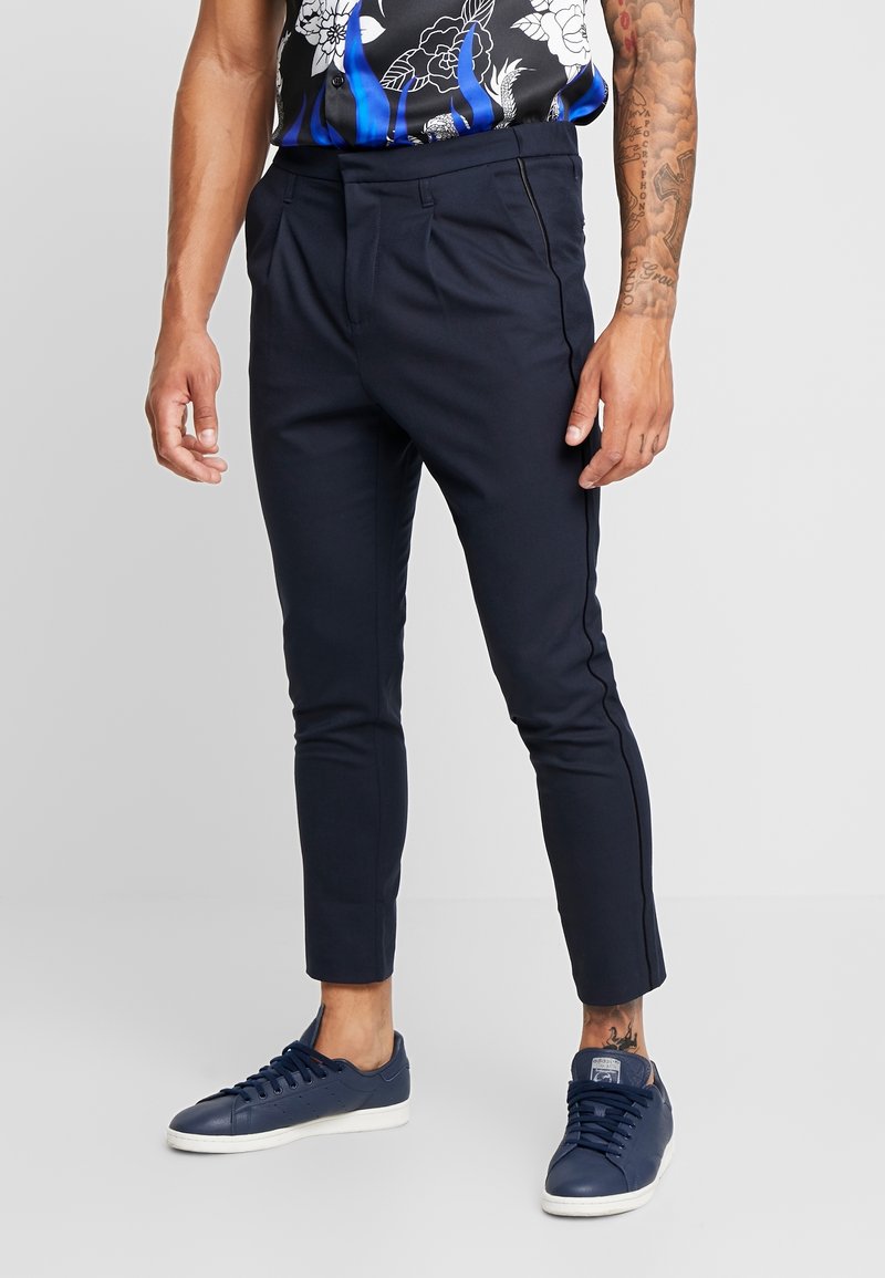 Homme portant un pantalon bleu marine foncé slim, des baskets bleu foncé et une chemise noire à manches courtes avec des motifs floraux bleus et blancs, se tenant devant un fond gris.