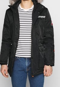 Parka negra con capucha forrada de piel, cierre frontal de cremallera y botones a presión, dos bolsillos laterales y detalle del logo en el pecho. Camisa a rayas debajo.