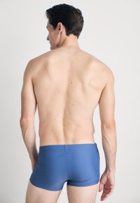 Shorts de bain pour hommes, bleus, avec une coupe ajustée, fabriqués dans un matériau lisse et extensible, sans motifs visibles ni détails matériels.