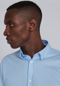 Chemise col bleu clair avec un design à boutons, texture lisse et boutons blancs, présentée en vue de profil.