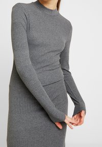 Abito lungo grigio a coste con maniche lunghe, scollatura alta e silhouette aderente. Presenta delle aperture ai polsini delle maniche per un tocco di dettaglio in più.