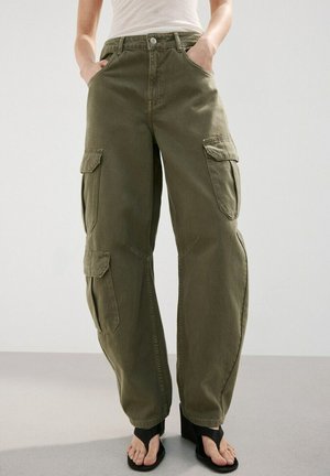 Persona che indossa pantaloni cargo color oliva a vita alta con numerose tasche e sandali neri con tacco e punta aperta, in piedi su uno sfondo semplice.