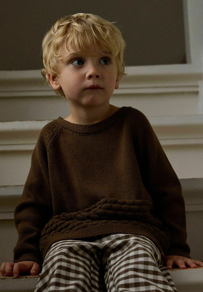 Jeune garçon blond portant un pull marron et un pantalon à carreaux, assis sur des marches blanches, regardant légèrement sur le côté.