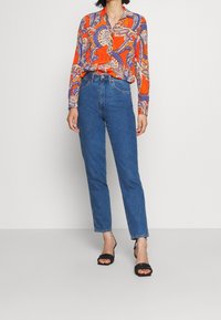 Färgglad blus med paisleymönster i orange och blått, kombinerad med högmidjade blå jeans och svarta klacksandaler.