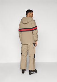 Ανοιχτό καφέ μπουφάν με κουκούλα, που διαθέτει μια κόκκινη και μαύρη οριζόντια ρίγα στη πλάτη. Συνδυασμένο με καβουράκια σε κρέμα με σχέδιο.