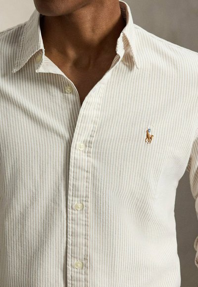 Homme portant une chemise à boutons rayée crème et beige avec un logo brodé de joueur de polo sur la poitrine gauche.