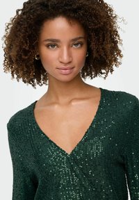 Zeleni top s svetlečimi odsevi in V-izrezom, teksturirano blago in oprijet kroj, ki vsebuje svetleče palčke v različnih odtenkih.