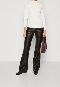 Maglione bianco a coste con orlo scallop abbinato a pantaloni neri in pelle a zampa d'elefante e una borsa bordeaux, su uno sfondo neutro.