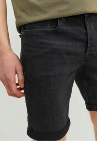 Hombre que lleva pantalones cortos de mezclilla negra arremangados y una camisa verde oliva, de pie con la mano relajada a un lado.