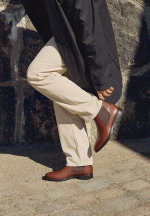 Une personne vêtue d’un manteau noir et d’un pantalon crème lève un pied pour montrer une bottine Chelsea en cuir marron, près d’un mur de pierre.