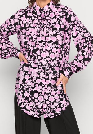 Žena nosící černou blůzu s dlouhými rukávy s růžovým květinovým potiskem a černé kalhoty, ruce opřené o boky.