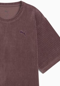 Brun fløjl t-shirt med ribbet tekstur, rund halsudskæring og et lilla broderet logo på brystet. Korte ærmer.