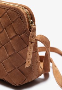 Torebka crossbody z zamszu w kolorze tan, o wplecionym wzorze, z złotym zamkiem błyskawicznym i regulowanymi paskami. Charakteryzuje się miękką, gładką powierzchnią i zaokrąglonymi krawędziami.